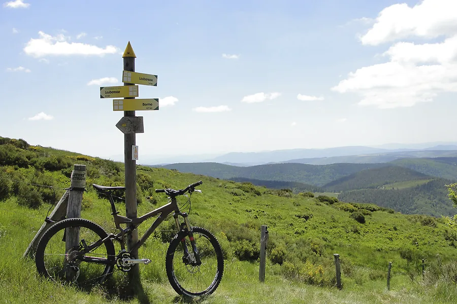 VTT au sommet d'un col devant un panneau de randonnée, panorama sur les monts d'Ardèche
