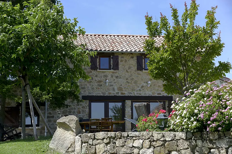 La façade en pierre de La Pradella et sa table en teck dans le jardin fleuri